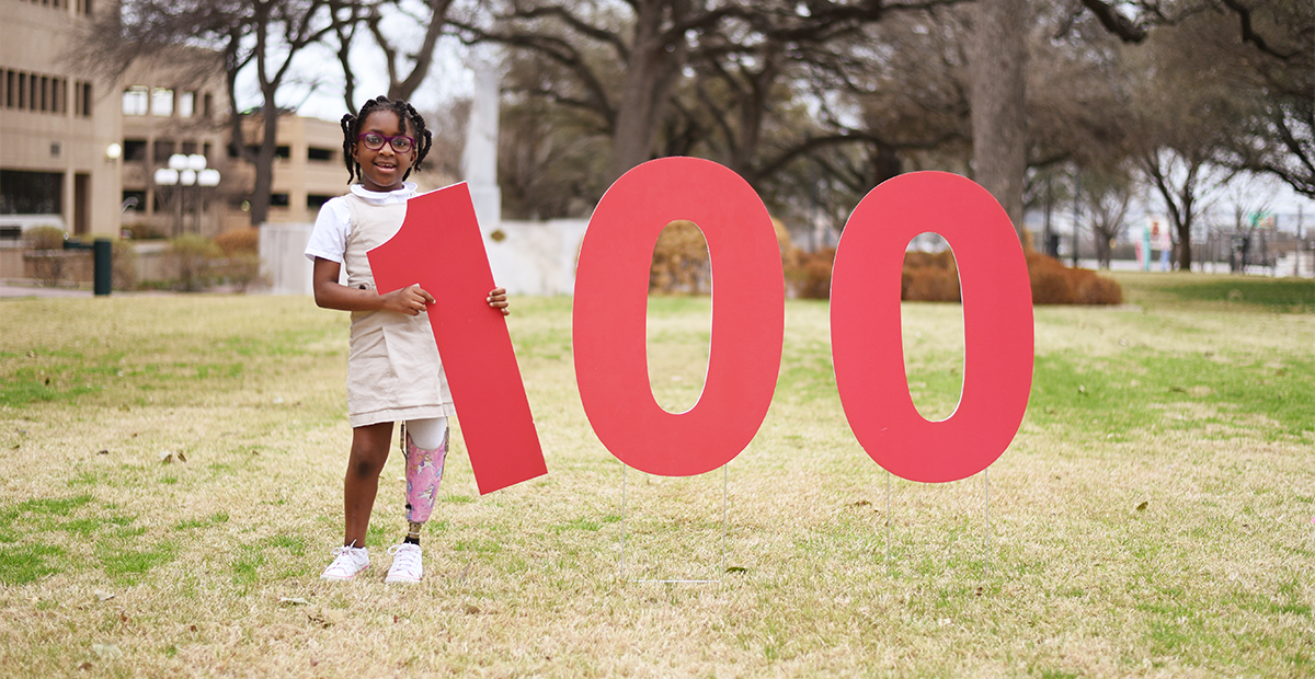 100. A little girl with a prosthetic leg holds up the number 100